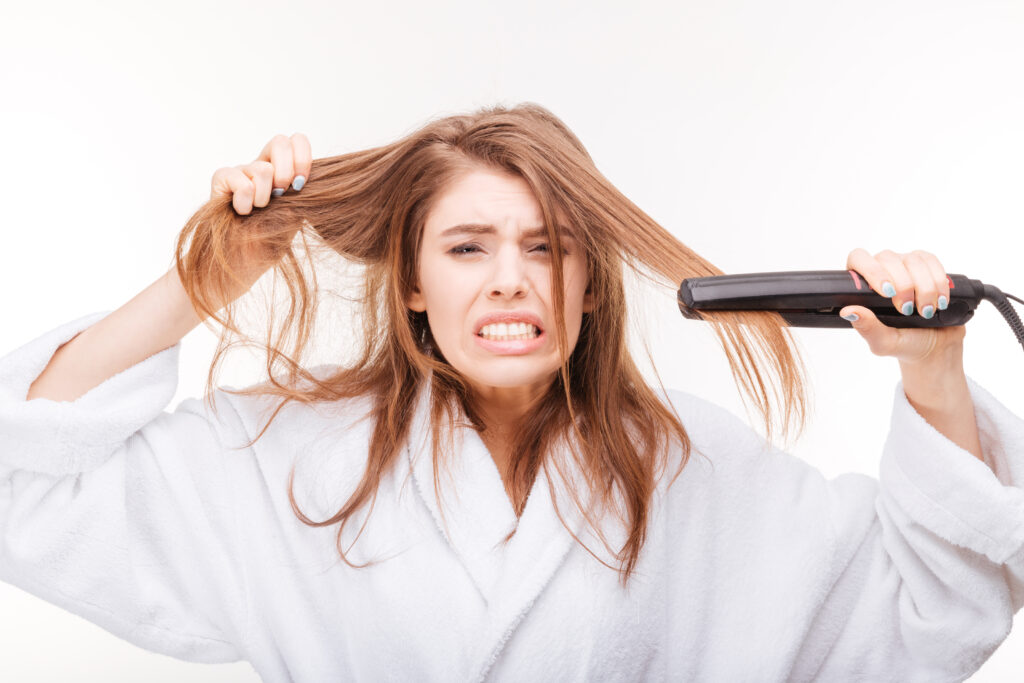 Angry irritated young woman straightening her hair using hair straightener over white background